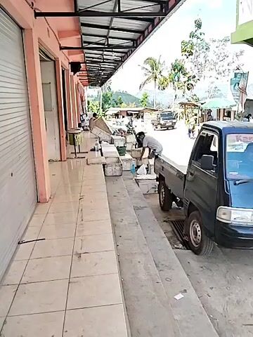 In a covered market area, a black pickup truck is parked next to a concrete walkway. A person is seen near the truck, appearing to be engaged in loading or unloading items from the truck bed, which is filled with white boxes. Another person is visible further down the walkway, near some open shopfronts with roll-down shutters. In the background, more stalls and structures suggest a bustling market environment, with trees and hilly terrain visible beyond. The sky is bright, indicating daytime.
