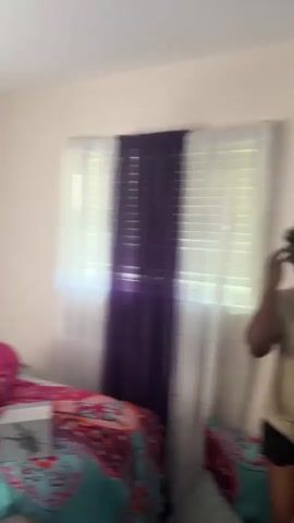 The video begins with a blurry shot of a bedroom with two beds, a window with purple and white curtains, and various decorations on the wall. The camera then focuses on a person wearing a pink bonnet, a dark sweatshirt, and dark pants, holding a vacuum cleaner. Next, a person wearing a tan shirt with the word "BLESSED" on it appears, holding a cardboard box and ripping paper inside. The camera zooms in on the person's face as they look down at the box.