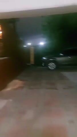 The video shows a dark outdoor scene at night. A wooden fence with lights on it runs along the left side of the frame. The ground is paved with interlocking stones. A dark-colored car is parked near a wooden gate in the background. The scene appears to be a driveway or courtyard. The video is shaky and the lighting is dim.
