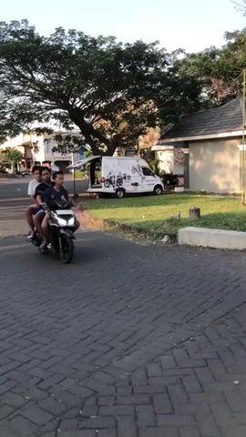 Three young men ride a scooter down a brick-paved road. The man in front is wearing a black t-shirt and blue shorts. The middle rider wears a gray t-shirt and dark shorts. The man in the back wears a dark t-shirt and shorts. All three wear short hair. The scooter is black. The men are smiling, and appear to be having fun. The scene is sunny, and the sky is clear blue. It is late afternoon or early evening. The setting appears to be a suburban or semi-rural area in Boro, East Java. There are trees and a building in the background. A white van with graphics on its side is parked in the background. The van has text that is not legible.