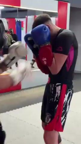 Two people are sparring in a gym. One person wears a black shirt, red and black shorts, and blue boxing gloves. The other wears a black shirt and shorts, white boxing gloves, and a black head guard. Both are throwing punches at each other. The gym has a mirror on the wall. There are other people in the gym.