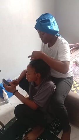 Two women are in a room. One is sitting on a bed and braiding the hair of the other woman, who is sitting on the floor, looking at her phone. The braider is wearing a blue satin bonnet, a white t-shirt, and dark pants. The other woman is wearing a grey t-shirt and dark pants. There is a white bottle and a blue container on the floor next to the woman with the phone. The floor has a black and white marble pattern. There is music playing in the background.
