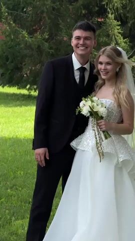 A young couple, a man and a woman, pose for a photo outdoors on a sunny day. The man is wearing a black suit with a white shirt and a black tie. The woman is wearing a white wedding dress with a strapless bodice adorned with sequins and a full skirt. She is holding a bouquet of white flowers. They are both smiling and looking at the camera. The background consists of green grass and tall pine trees. The video is short, showing the couple in the same pose for the duration.