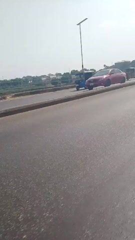 The video shows a roadway with various vehicles and a pedestrian. At the beginning of the video, a motorcycle and an auto rickshaw are visible in the distance. A black car then passes by the camera. A pedestrian dressed in white can be seen walking on the side of the road. The background includes buildings and trees in the distance.  The roadway appears to be a bridge over a body of water. There is a light pole near the edge of the road.