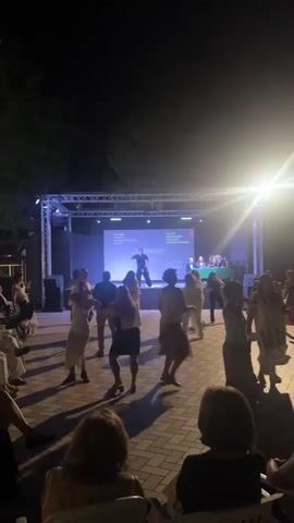 A man is dancing on a stage at night. He is wearing a black t-shirt and black pants. There is a large screen behind him with text on it. Several people are dancing in front of the stage. Some people are sitting in the foreground watching the performance. The event appears to be outdoors.