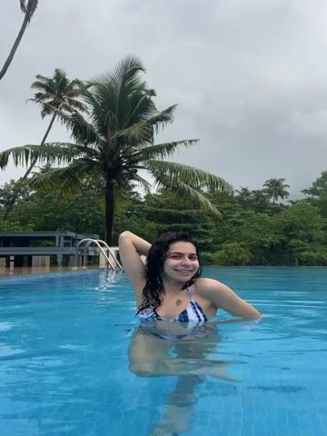 The video shows a woman in a swimming pool. She is wearing a blue and white tie-dye bikini and has curly brown hair. Her left arm is raised, and she is smiling at the camera. Behind her, there is a lush green landscape with palm trees, and the sky is overcast. The water in the pool is a vibrant blue. The camera is positioned at water level, capturing the woman from the waist up. The video appears to be a short clip or a series of short clips stitched together.