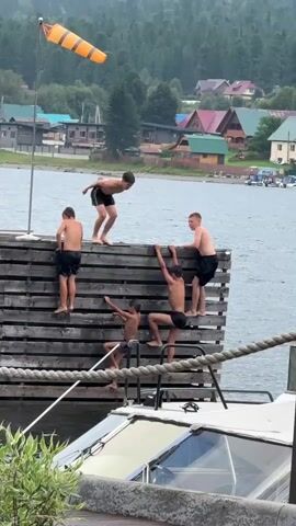 The video shows a group of young boys on a wooden dock next to a body of water. Several boys are climbing on the side of the dock, using the horizontal planks for support. One boy is in the process of jumping or diving into the water from the top of the dock. Another boy is assisting him from below, holding onto his leg. In the background, there is a shoreline with houses and trees, and a windsock is visible on a pole. A boat is docked below the boys. The overall scene depicts a recreational activity involving climbing and jumping into the water.