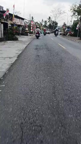 The video shows a first-person view of riding a motorcycle on a paved road. Several other motorcycles and a car are visible ahead on the road. The road has a yellow line marking the center and appears to be in a rural or suburban area, with buildings, trees, and flags lining the sides. The weather appears overcast. The motorcyclist is wearing a helmet.