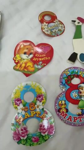 The video shows a flat lay of several decorative items on a white, textured surface. The items are of various shapes and sizes, and appear to be vintage or retro in style, possibly intended for decoration or as small gifts.

In the upper left quadrant of the frame is a small, red and gold decorative item shaped like the number 8, with floral embellishments. Next to it, slightly below and to the left, is a red heart-shaped decoration featuring a cartoonish orange kitten with a yellow bow around its neck. Inside the heart, two pink smaller hearts are visible, and the text "БУДЕМ ДРУЖИТЬ?" (Let's be friends?) is written in pink.

To the right of the kitten heart, a stylized, cutout figure of a bird or peacock is visible. It is predominantly white with a green neck, a pink head accent, and colorful accents on its body and tail feathers, including pink, blue, and yellow circles.

Below the kitten heart and to the left, there are two circular decorations stacked vertically, both shaped like the number 8 and adorned with images of pink roses and green leaves. The top circle has a blue background and yellow floral accents, with the text "8 МАРТА" (March 8) in the center. The bottom circle also has "8 МАРТА" written on it, with more prominent pink roses and green foliage.

To the right of the stacked circles, and partially overlapping the bird figure, is another large, red number 8 shaped decoration. This one features cartoon characters, including a child wearing a blue hat and green clothing, and a small bee. The text "8 МАРТА" is also visible on this decoration.

The lighting is soft and appears to be coming from above, casting slight shadows. The white surface has a subtle texture, possibly fabric or paper. The overall impression is one of a collection of festive, possibly celebratory, decorative items.