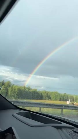 The video shows a view from inside a vehicle, looking through the windshield. Outside, a double rainbow is visible in a cloudy sky. The landscape features a road, trees, and fields. The road appears wet, suggesting recent rain. In the distance, there are some structures that might be buildings or power lines. The camera pans slightly, offering different perspectives of the rainbow and the surrounding scenery.