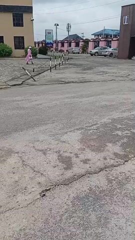 The video shows a street scene with several buildings and people. In the foreground, the ground is made of cracked asphalt. A line of white-capped bollards runs along the edge of the road. In the middle ground, a person in a pink dress walks away from the camera. Behind them, a light-colored building with dark windows stands. Further back, several cars are parked. In the background, there are more buildings, including a large building with a prominent red cross on its facade, suggesting it might be a church or a religious institution. People are seen walking near this building. The sky is overcast. The video appears to be filmed from a moving vehicle or by someone walking.