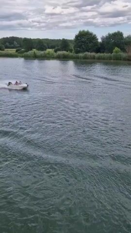 A white boat with four people aboard travels from left to right across a calm body of water. The boat leaves a white wake behind it. On the far bank, green trees and vegetation line the shore. The sky is overcast with a mix of blue and gray clouds.