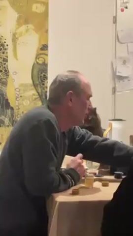 Here is a description of the video:

The video shows a man with short, graying hair wearing a gray long-sleeve shirt, sitting at a table with a tan tablecloth. He is gesturing with his hands and talking. On the table, there are small glasses filled with a golden liquid, poker chips, and a bottle of alcohol. In the background, behind the man, there is a fluffy brown cat sitting on something that is not visible. There's an abstract painting on the wall to the left, and a white cabinet with papers attached to the door on the right. The lighting in the room appears to be standard indoor lighting. Another person is partially visible to the right.