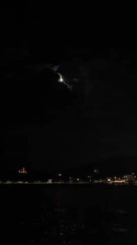 The video shows a night scene with a partially obscured moon in the sky. Below the moon, there is a dark body of water reflecting the moonlight. In the distance, a faint outline of a city or town is visible with scattered lights. A building that appears to be a church or monument with a dome-like structure is illuminated on the left side of the frame. The overall atmosphere is dark and tranquil.