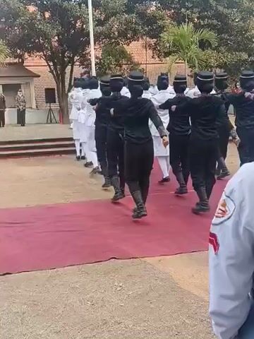 A group of individuals in matching uniforms, some in black and others in white, are marching in formation outdoors on a dirt and gravel area with a red carpet laid out. The individuals in black are at the front, facing away from the camera, and appear to be performing a synchronized movement with their arms. Behind them, a line of individuals in white uniforms are also marching. In the background, there is a brick building, trees, and other people watching. The overall scene suggests a formal event or ceremony.