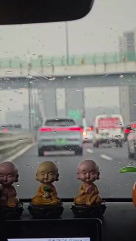 The video shows a close-up view from inside a car, looking out the windshield at a highway. Raindrops are visible on the windshield. In the foreground, three small figurines of smiling Buddhist monks are placed on the dashboard. They are in a seated position, with their hands clasped. The middle monk is holding a green lotus flower. The road ahead is filled with traffic, including cars and a white van with red lettering. The sky is overcast, and the overall lighting suggests it is daytime, possibly during a rainy or foggy weather condition.