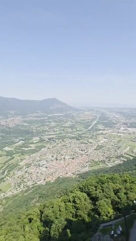 The camera pans across a broad, sunlit landscape from a high vantage point. In the foreground, lush green trees form a dense border, while a tall, gray stone structure appears on the left side of the frame. Beyond the trees, a sprawling town or city is visible, nestled among fields and patches of green. Roads and highways wind through the landscape, connecting different areas. Further out, a range of mountains rises against the clear blue sky, creating a picturesque backdrop. The scene evokes a sense of vastness and serenity.