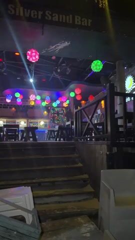 The video captures an interior night scene of a bar identified as "Silver Sand Bar" by a sign overhead. The space is dimly lit by multicolored disco lights, including numerous spherical, textured lanterns suspended from the ceiling in various colors like pink, yellow, blue, and green. Laser-like light beams also cut through the darkness. In the background, a bar counter with stools is visible. To the right, a raised seating area with wooden railings features tables and chairs. Stairs with yellow safety strips descend from this area towards the camera. In the foreground at the bottom of the stairs, several white plastic chairs and a wooden crate appear overturned or haphazardly placed. A person with dark hair in a white top is briefly visible near the bar on the left side of the frame. Another person with light-colored hair, wearing a light-colored shirt, is seen sitting at a table in the raised area on the right. The camera subtly pans slightly to the right.