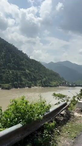 The video pans across a wide, muddy river flowing through a valley. Lush green, tree-covered mountains rise steeply on either side of the river. The sky is partly cloudy with a bright sun. In the foreground, a concrete barrier runs along the edge of a road, separating it from the river. A single plastic water bottle is tied to a thin metal pole protruding from the barrier. The camera movement is smooth, capturing the expansive natural scenery.