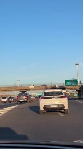 The video shows a view from a car driving on a highway with moderate traffic. The sky is clear blue. In the foreground, there is a white hatchback with license plate AE 627 YF. Other cars including an SUV and a grey sedan are visible. An overpass crosses the highway with a sign indicating "CALLE THAMES" and other street names in 350 meters. There are people walking on the overpass. Streetlights are visible. The camera is stable and appears to be mounted inside the car.