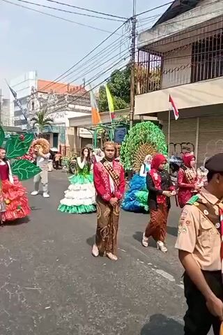 The video shows a parade on a sunny day. Several people are walking in the street, some wearing elaborate costumes. In the foreground, a young person wearing a red and gold traditional outfit and a gold headpiece stands facing the camera. To their left, a person in a peacock-like costume with green and blue feathers is visible. Further left, a person in a green and white tiered dress walks alongside a person in a lion costume. On the far left, a person in a red and gold dress with large green leaf-like structures attached to their shoulders is visible. Several other participants in traditional clothing and costumes are present in the background. Buildings line the street, and flags are visible.
