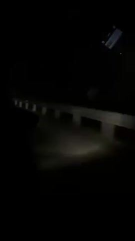 A vehicle moves along a dark, winding road, illuminated by its headlights. The road is bordered by a white guardrail on one side. The vehicle's dashboard is partially visible in the frame. The environment appears to be rural or mountainous, with no other vehicles or significant landmarks present. The video seems to be taken from the driver's perspective.