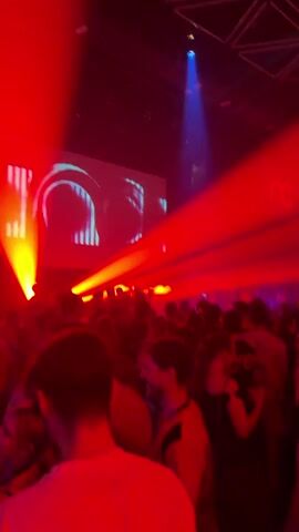 The video shows a crowded indoor venue, likely a nightclub or concert hall. A large screen at the back displays abstract, glowing red and white graphics, resembling metallic arches or pathways. Powerful red laser beams cut through the haze, illuminating the crowd and the screen. The audience, a mix of young adults, appears to be dancing and enjoying the music. Some individuals are holding handheld fans, one pink and white, another red. The lighting is predominantly red and white, with a blue spotlight visible in the upper right corner. The atmosphere is energetic and festive.