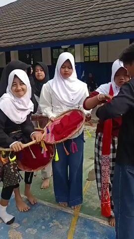 The video shows a group of young women outdoors, dressed in hijabs and traditional clothing. They appear to be participating in a cultural event or performance. Two women in the foreground are holding woven circular frames adorned with colorful tassels. One woman is wearing a black jacket over a patterned skirt and white hijab, and the other is wearing a white shirt and blue pants with a white hijab. Another woman in the foreground, wearing a black hijab and white top, is holding a woven tray filled with fabrics. In the background, other women in similar attire are visible. The environment appears to be a courtyard or open space with buildings in the background. There are some decorative elements and possibly musical instruments present. The interactions suggest a coordinated activity, possibly a dance or a ceremony.