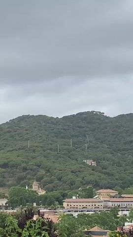 This video shows a panoramic view of a landscape with a large, tree-covered hill dominating the background under a cloudy, grey sky. In the foreground and midground, a town is visible with various buildings, including residential structures and what appears to be a historical building or church with a bell tower on the left side of the frame. Power lines and utility poles can be seen ascending the forested hillside. The camera slowly pans from left to right, revealing more of the town and the expanse of the hill. The trees on the hill appear to be evergreens, likely pines, given the location context of Vilassar De Dalt, Spain. The weather is overcast, suggesting a cloudy day. No people or visible text are present.