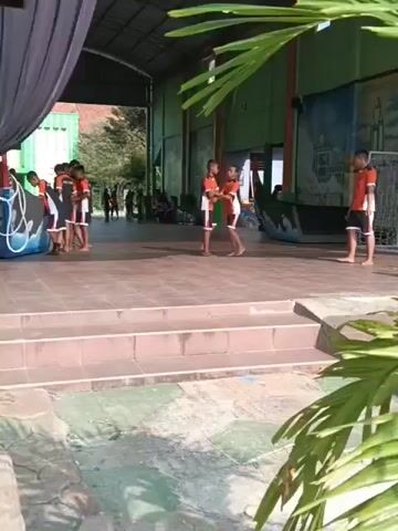 The video shows an outdoor covered area with a tiled floor and steps leading up to a stage-like platform. Several young people wearing orange shirts and dark shorts are on the platform. Some are standing and talking, while others appear to be playing or practicing. There is a soccer goal visible in the background. In the foreground, there are large green leaves obscuring some of the view. The lighting suggests it is daytime.