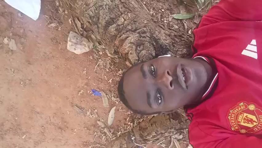 A young man with dark skin is lying on the ground. He is wearing a red Manchester United soccer jersey. He has white earbuds in his ears. The ground he is lying on is a mix of dirt and roots from a tree. There is some trash scattered around him. He is looking directly at the camera and talking.