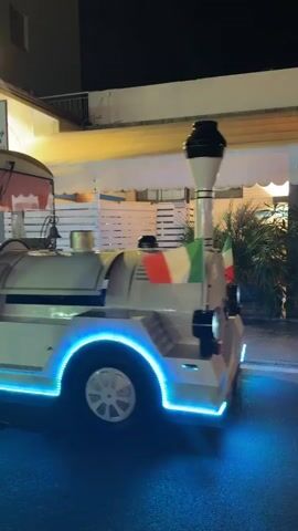 A white tourist train with blue LED lights moves along a street at night. The train has a sign that reads "DOLCIARIA Dalba" and "dal 1930". The train appears to be decorated with orange curtains and has an Italian flag. People are visible inside the train, including a driver. In the background, there are buildings and lights, suggesting a commercial or entertainment area. The video is filmed from a side angle, following the train.