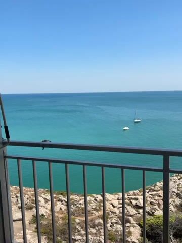 The video shows a panoramic view from a balcony overlooking a coastal landscape. In the foreground, a metal railing with vertical bars runs across the bottom of the frame, partially obscuring the view. Beyond the railing, a rocky cliff face with sparse vegetation is visible, dropping down towards the sea. The sea itself is a vibrant turquoise color and stretches to the horizon, meeting a clear, bright blue sky. Several sailboats are scattered across the water, their white sails and hulls standing out against the blue. In the middle ground, a section of buildings with pink and white facades and visible balconies can be seen nestled on the coast. The overall scene depicts a sunny day at a seaside location, likely a resort or a coastal town.
