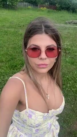 A young woman with blonde hair and pink sunglasses smiles at the camera while standing in a grassy field. She is wearing a light yellow dress with a floral pattern and a gold cross necklace. The background shows trees, a wooden fence, and a cloudy blue sky. The woman is likely in a rural area, possibly Russia, given the location context. The sun is shining, indicating it is daytime. The overall mood appears cheerful and relaxed. There is no visible text in the media file.