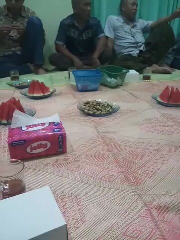 Here is a concise description of the video:

The video shows a group of men sitting on the floor around a low table. The men are dressed in casual clothing. There are plates of watermelon slices and a bowl of peanuts on the table. A tissue box and a decorative table are also visible. The camera pans across the scene, showing the men talking and eating. The room has a light green wall and a pink patterned mat.