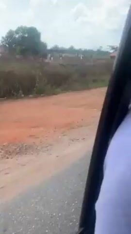 The video shows a view from inside a moving vehicle. The car is traveling on a paved road with a dirt shoulder. In the distance, there are small buildings and sparse vegetation. A sign reading "Welcome To (Unreadable)" is also visible. The sky is partially cloudy.  A person's shoulder and arm are visible in the lower right portion of the frame, suggesting the point of view is from a passenger in the vehicle.