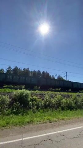 A train with open-top cargo cars is traveling on tracks. The train cars have the word NOVOTRANS written on them. The train is surrounded by greenery and trees. The sky is clear and the sun is shining brightly. The video is filmed from the perspective of someone in a vehicle. The vehicle's dashboard and steering wheel are visible in some frames. There is a side mirror reflecting the road ahead. The vehicle is driving on a paved road. The road is cracked in some places. There are power lines running along the train tracks. The train is moving away from the camera.