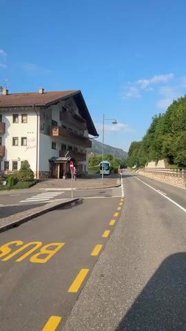 The video shows a view from inside a vehicle driving down a road on a clear day. A white building with a brown roof is visible on the left side of the road, with balconies and windows. A bus stop sign and a bus stop shelter are located in front of the building. Further down the road, a white bus is parked. On the right side of the road, trees line the hillside. The road has a crosswalk and traffic lights. The vehicle appears to be moving at a moderate speed, and the sun is casting shadows.