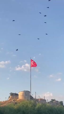The video begins with a static shot of a red flag with a white crescent and star, the flag of Turkey, flying on a flagpole. The flagpole is situated on a hilltop in front of a circular concrete structure. Several dark, bird-like objects are scattered across the clear blue sky. As the video progresses, more of these objects appear, and some are seen in motion, suggesting they are birds in flight. The camera remains stationary throughout the short clip, focusing on the flag and the surrounding sky. The environment appears to be outdoors with green foliage visible at the bottom of the frame. The lighting suggests it is daytime, possibly late afternoon given the warm tone of the sky. No people are visible in the video.