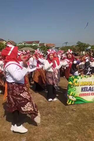 A group of women wearing red headscarves and white shirts are standing in a field, some of them appear to be clapping or performing a dance routine. They are also wearing patterned skirts. Behind them, there is a large green banner with yellow text that is partially visible. The banner appears to say "MI NURUL KELAS CERDAS." The sky is clear and blue, and there are buildings visible in the background. The ground is covered in dry grass.