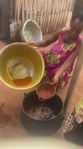 The video shows a woman in a pink dress pouring liquid through a small metal strainer into a large black pot. The woman holds a large silver pot high over the black pot and is tipping it so the liquid, which is orange, will flow into the strainer. A small amount of orange liquid remains in the silver pot. The strainer sits on top of the black pot. The black pot appears to contain a dark liquid or material. A second person, wearing a leopard print shirt, stands partially in the right side of the frame. Chickens are visible behind the woman. The scene takes place outside on dirt ground. There are bamboo poles and other pots in the background.