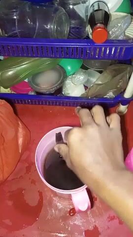 A person's hands stir a dark liquid in a pink mug on a red surface that appears wet and stained. Behind the mug, a purple plastic shelf unit holds various containers, including glass jars, plastic containers, and bags of contents. To the left of the mug is a red plastic bag. The lighting is dim and the scene appears to be indoors, possibly in a kitchen or storage area.