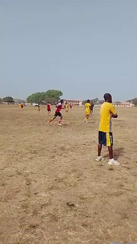 The video shows a soccer game in progress on a large, dry field with sparse grass. Several players wearing red jerseys compete against players wearing yellow jerseys. The players are spread across the field, and the ball is in the air at one point. A man wearing a yellow and blue uniform stands near the camera, watching the game. In the background, a building with multiple windows is visible. The sky is a pale blue.