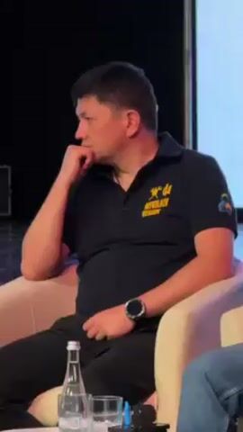 Here is a detailed description of the video:
The video shows a man sitting in a light-colored armchair. He is wearing a dark blue polo shirt with yellow text on it, black pants, and a wristwatch. The text on his shirt reads "Mykolaiv REGION." His right hand is resting on his knee, while his left hand is supporting his chin. His face is turned to the left, and he appears to be listening intently. A partially visible person is seated next to him, wearing blue jeans. A sealed bottle of water and a clear glass are visible on a small surface next to the man. The backdrop is a dark-colored wall.