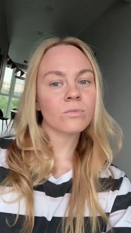 The video shows a close-up shot of a blonde woman with light skin, wearing a black and white striped t-shirt. She is looking at the camera with a neutral expression. Her hair is long and wavy, framing her face. The background appears to be an indoor setting, with a window visible on the left side of the frame. The lighting suggests it is daytime. The woman's face shows some redness on her cheeks. She blinks and slightly shifts her gaze during the short clip.