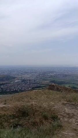 The video presents a panoramic view from a hilltop overlooking a city and surrounding landscape in Koy Tash, Kyrgyzstan. The scene transitions between different perspectives, revealing the expansive terrain.

Initially, the view showcases a city nestled in a valley, with a scattering of buildings and roads visible. The foreground is dominated by dry, brownish grass and sparse vegetation, suggesting a somewhat arid environment. The sky is overcast with soft, diffused light, indicating it might be morning or late afternoon.

As the video progresses, the camera pans to reveal more of the distant landscape, characterized by rolling hills and vast agricultural fields. The weather appears to be changing, with darker, more ominous clouds gathering on the horizon, hinting at potential rain or a storm. This shift in the sky adds a dramatic element to the scene.

Further on, the view expands to encompass a wider vista, with mountains visible in the far distance, partially obscured by haze. The city appears to be spread out across the plain, with a network of roads connecting different areas. The changing cloud formations continue to be a prominent feature, with patches of clearer sky interspersed with darker clouds.

The overall impression is one of a vast and somewhat rugged natural landscape interacting with human settlement. The changing weather and the expansive views evoke a sense of openness and perhaps a touch of melancholy or awe. There is no visible text in the video. The time of day is difficult to pinpoint precisely due to the overcast conditions but appears to be during daylight hours.