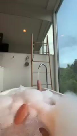 Here is a concise description of the video:

The video shows a person relaxing in a bubble bath next to a large window. The bathtub is white and filled with fluffy white bubbles. A silver faucet is running, adding more water to the bath. Visible in the bath are the person's legs and feet. A wooden ladder rests against the wall next to the bathtub. Through the window, green trees and a cloudy sky are visible. The room has a modern design.