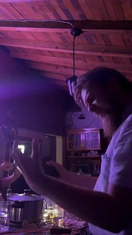 A man with a beard and glasses sings into a microphone, gesturing with his hands. He is wearing a light-colored shirt. The background is dimly lit with purple and red lights. Other people are visible in the background, one of whom is playing a guitar. There is a bottle of "DEVIN" water on a table in front of the man. The room has a wooden ceiling.