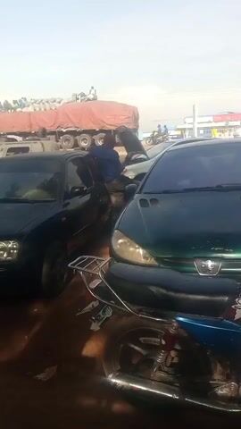 The video shows an outdoor scene, possibly a parking area or roadside in an urban environment. Several vehicles are visible: a dark green Peugeot sedan partially obscured by a blue and red Suzuki motorcycle in the foreground, a dark-colored sedan to the left, and other cars and a large truck with a red tarp in the background. A man in a blue shirt is standing by a car with its hood open. Another man wearing a gray shirt and an orange cap walks past the camera, moving from left to right, across the front of the green Peugeot. There are buildings and utility poles visible in the background, under a slightly hazy sky. The ground appears to be dirt or gravel. There is ambient noise that includes voices speaking.