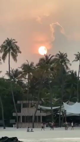 The scene is a beach with a dusky orange sky, presumably at sunset. The sun, appearing as a bright orange orb, is partially obscured by clouds. Silhouetted palm trees line the beach, adding to the tropical ambiance. In the foreground, there is a wooden structure resembling a bar or stage. Several people are visible on the beach, some standing and talking, others walking around. A white tent-like structure is also present to the right. The sand appears white and fine. The overall scene suggests a relaxed, vacation-like atmosphere.